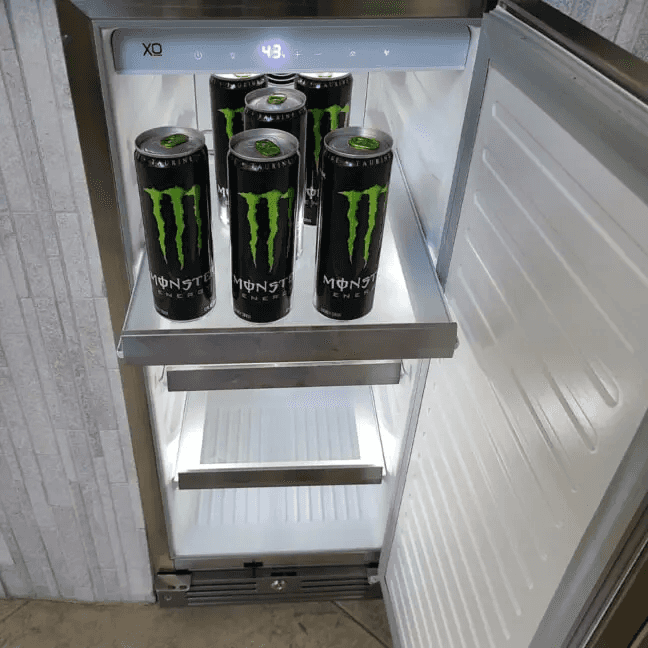XO 15 Inch Outdoor Undercounter Refrigerator interior view with Monster energy drinks on top shelf, showing LED lighting and temperature display.