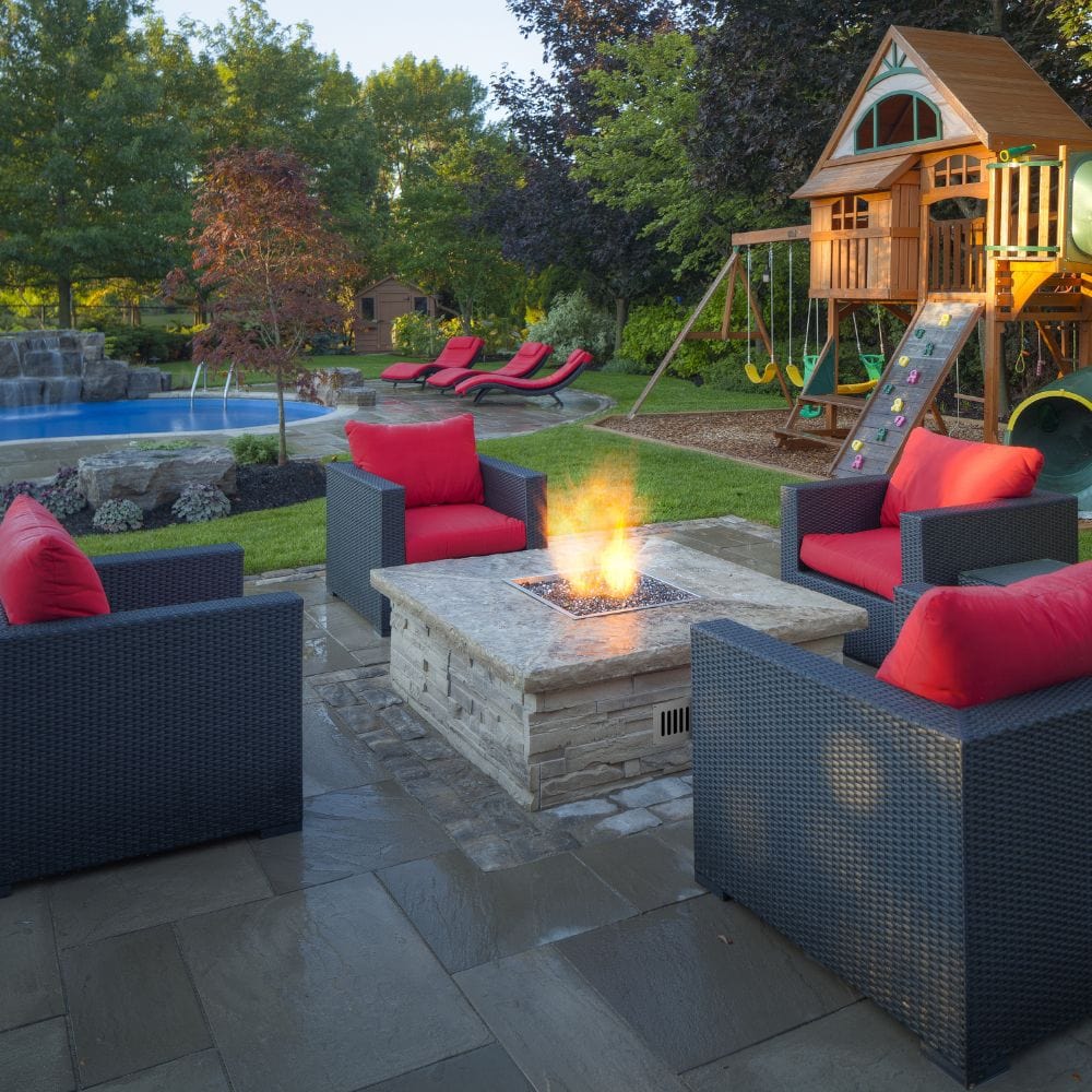 Backyard patio setup featuring Firegear DIY Fire Pit Kit - Square in a stone base surrounded by wicker chairs and red cushions.