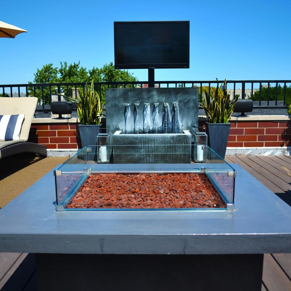 Close-up of Firegear DIY Fire Pit Kit - Square with lava rock and glass wind guard, placed on a rooftop patio with water feature.