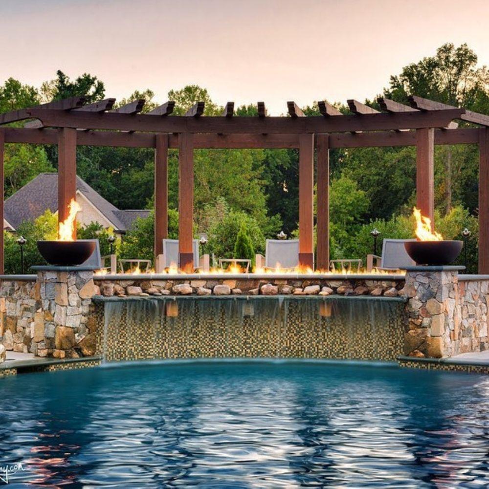 Firegear Sanctuary 30-Inch Round Concrete Gas Fire Bowls on stone pillars beside pool waterfall under wooden pergola.