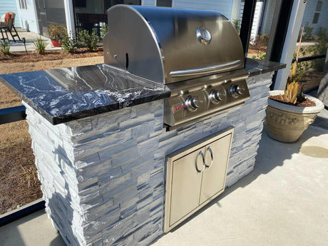 Compact stacked stone grill island with a built-in Blaze 25 inch gas grill, double access doors and a black countertop on a patio