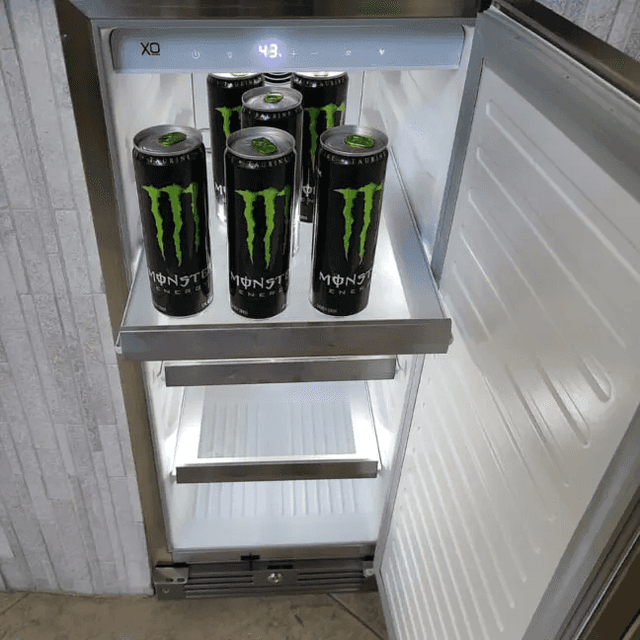 XO 15 Inch Outdoor Undercounter Refrigerator interior view with Monster energy drinks on top shelf, showing LED lighting and temperature display.